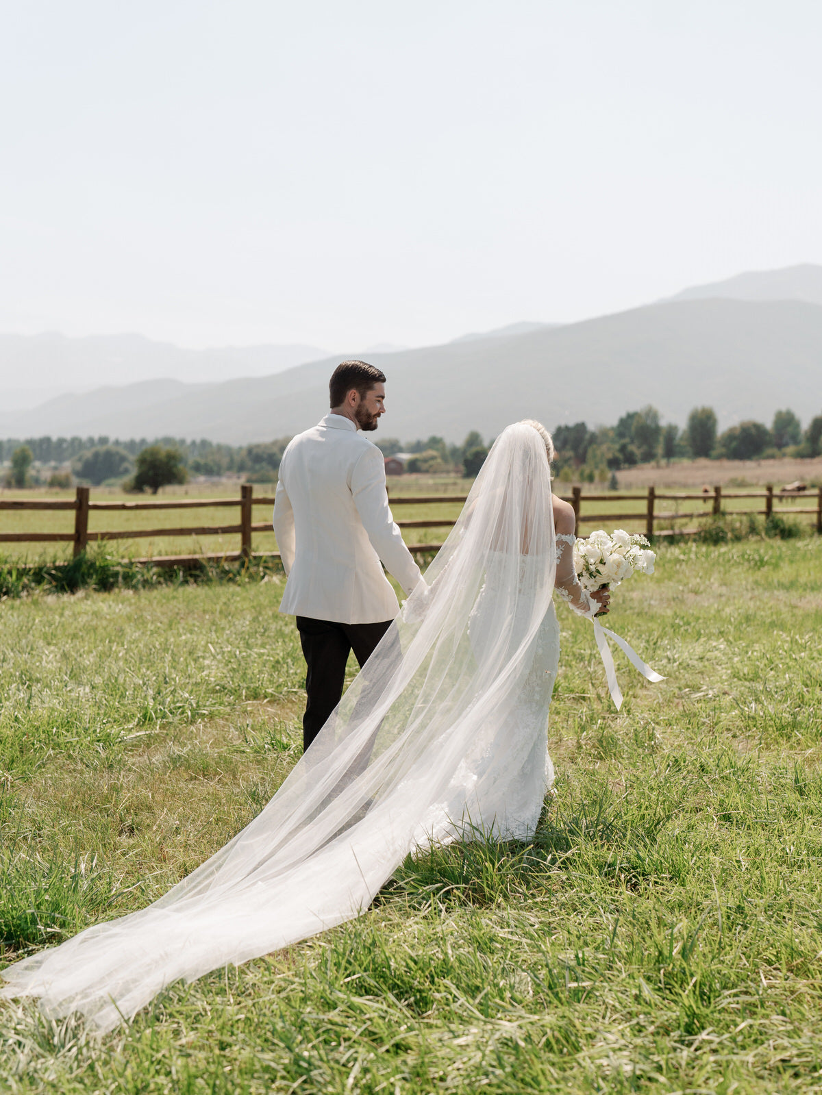bride and groom in wedding attire standing in a grassy field with bride in extra long royal length 150 inch bridal veil in off white