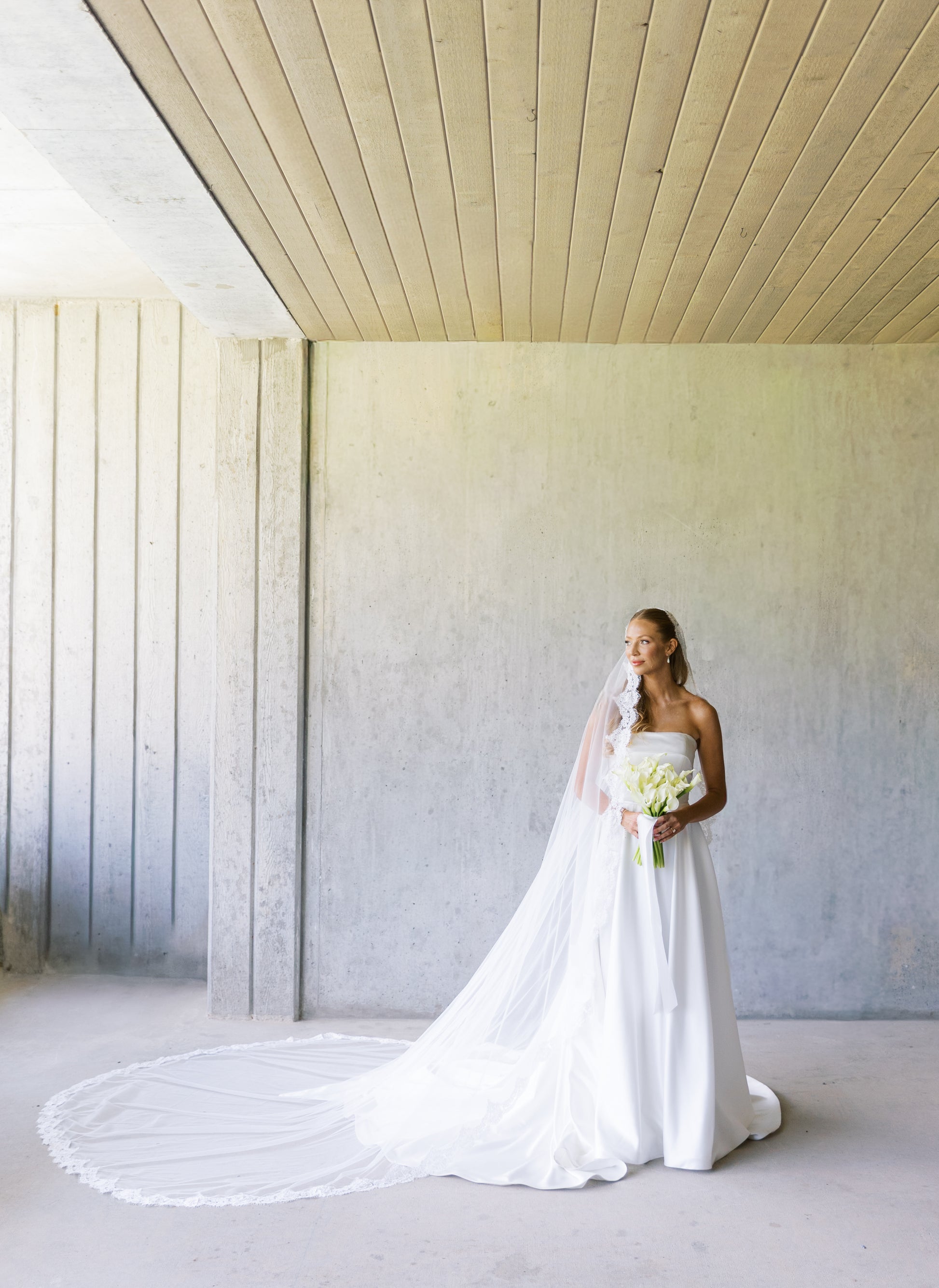 Bride in a timeless and clean wedding dress standing in a minimalistic room with long mantilla wedding veil royal length on top of Half up half down hairstyle