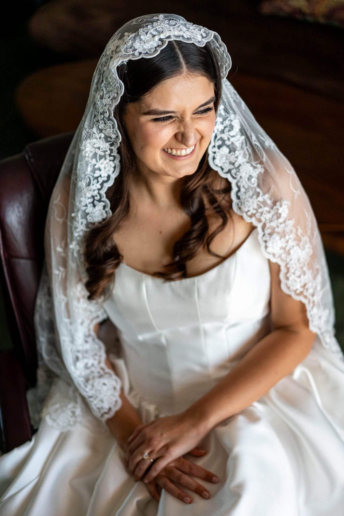 Woman in a white wedding dress with a lace mantilla veil over loose downdo, sitting indoors in cat eye neck gown