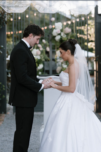 strapless ballgown satin gown with fingertip white raw edge veil under high curled updo as bride stands at alter