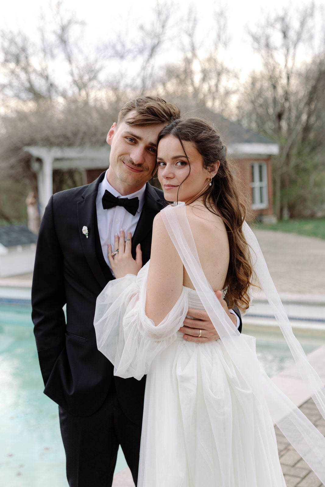 groom in a tuxedo and bride in a white off shoulder dress wearing narrow neck scarf standing by a pool.
