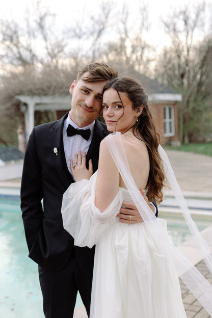 groom in a tuxedo and bride in a white off shoulder dress wearing narrow neck scarf standing by a pool.