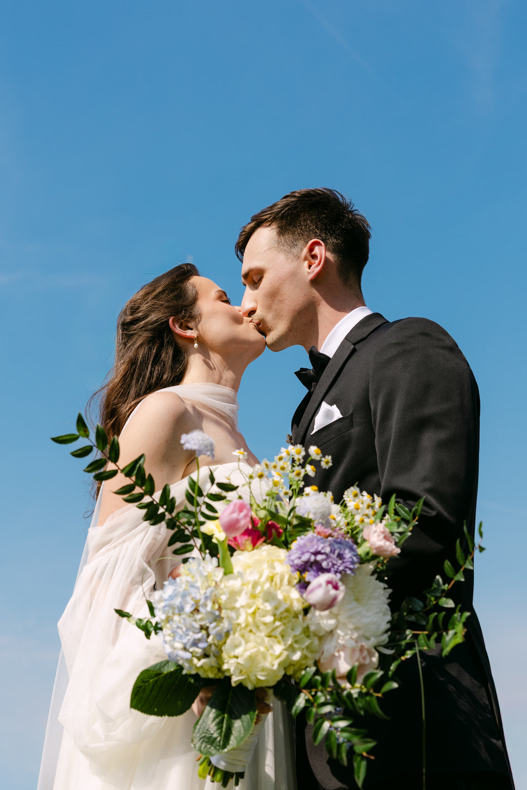 Couple kissing with bride in long narrow bridal neck scarf choker and a bouquet of flowers against a clear blue sky