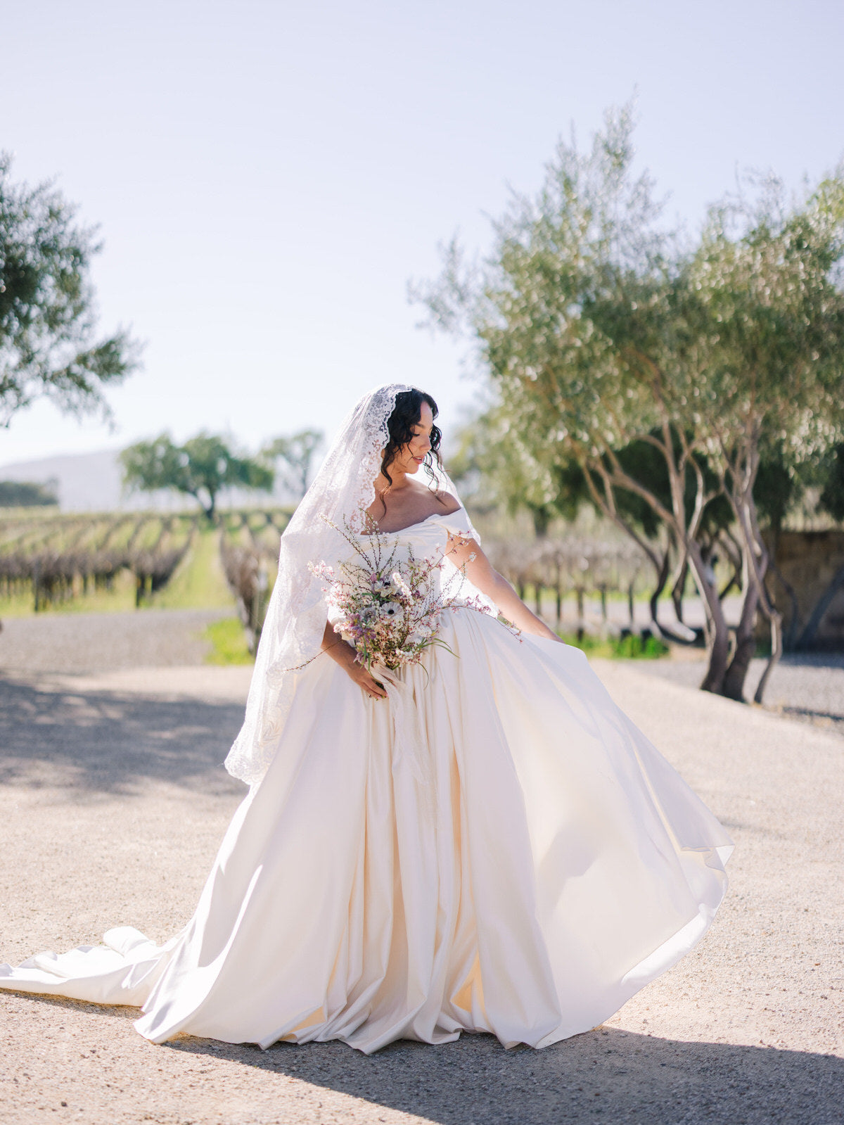 Bride in a white off shoulder ballgown wedding dress with a fingertip lace mantilla veil standing in California winery
