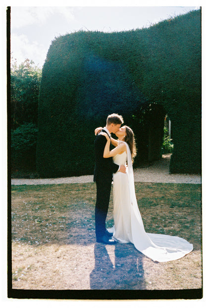 engaged couple embracing with trailing neck scarf  in a garden setting with large hedges.