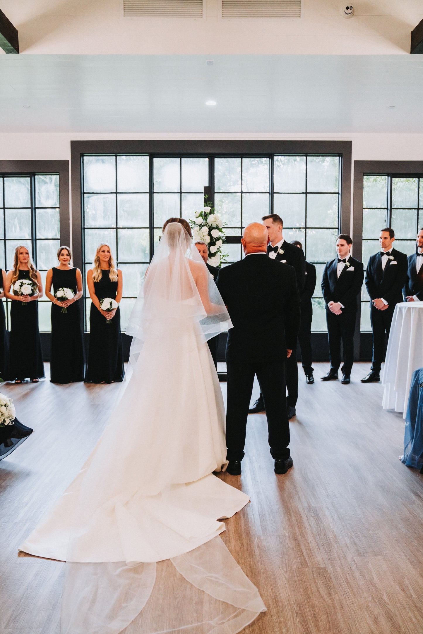 black tie Wedding ceremony with a bride walking down the aisle in drop two tier wedding veil with horsehair ribbon trim and bridesmaids in black long gowns