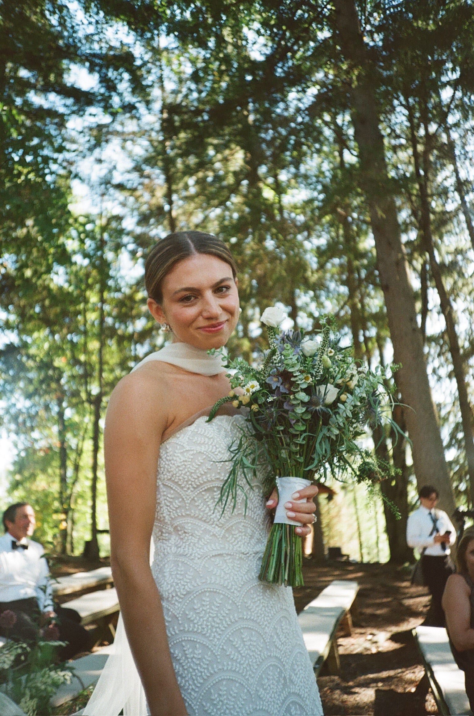 Woman in a lace wedding dress and narrow bridal scarf holding a bouquet of wildflowers outdoors with trees in the background