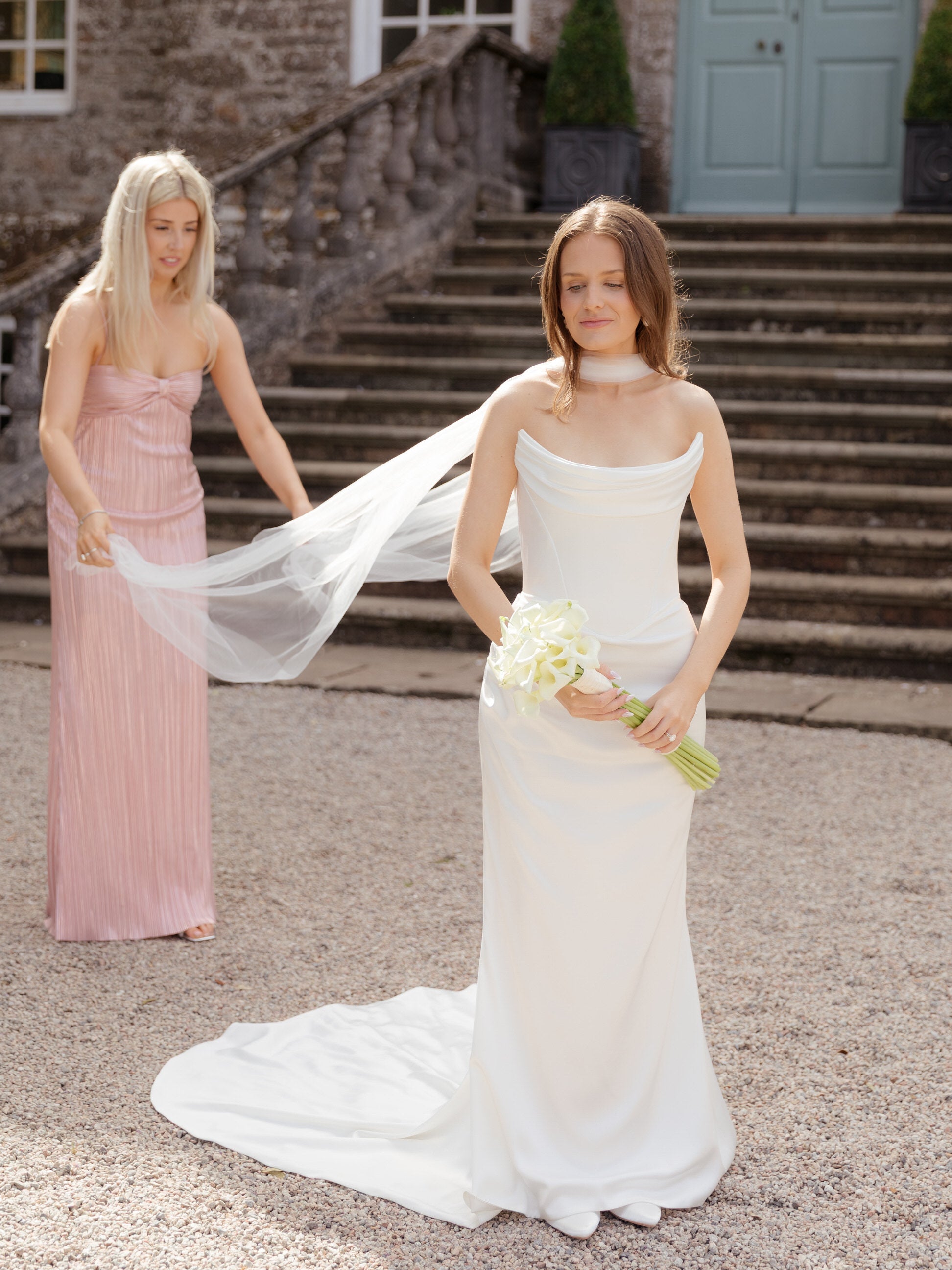 bride in a cat eye neckline wedding dress holding a lily bouquet, with bridesmaid in a pink dress helping her put on narrow bridal neck scarf