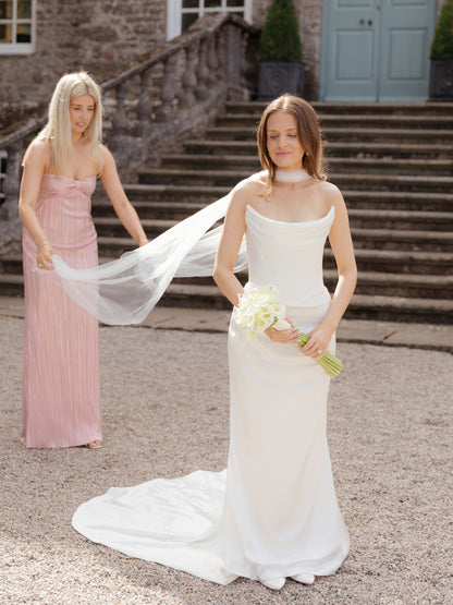 bride in a cat eye neckline wedding dress holding a lily bouquet, with bridesmaid in a pink dress helping her put on narrow bridal neck scarf