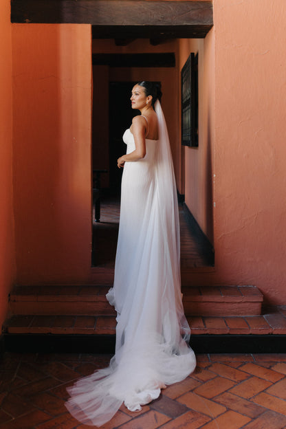 A bride in a spaghetti strap white wedding gown with a long English net veil stands in profile in a rustic corridor with terracotta walls and brick flooring.