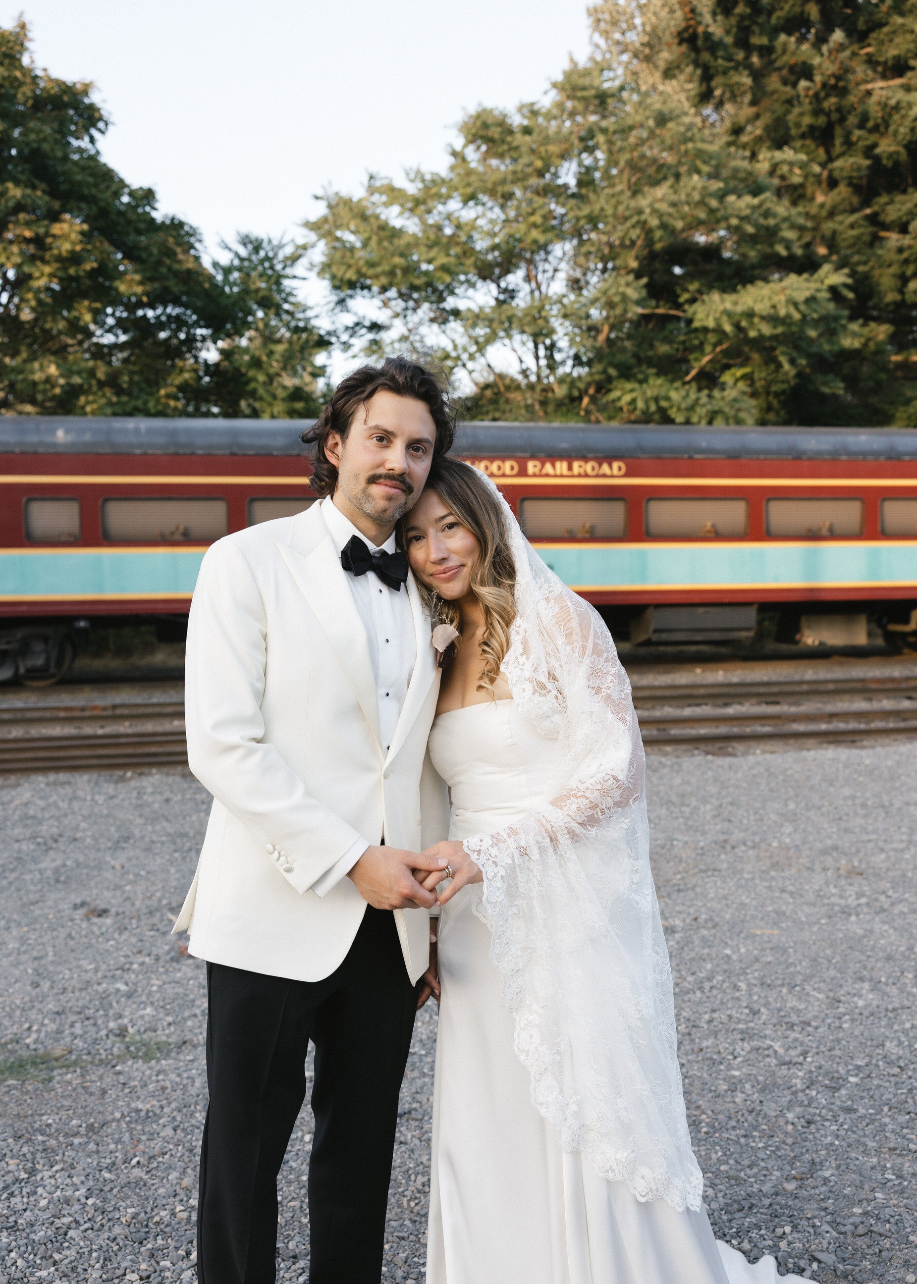 Bride and groom in wedding attire standing in front of a vintage train. bride wearing ballet length chantilly lace veil from one blushing bride