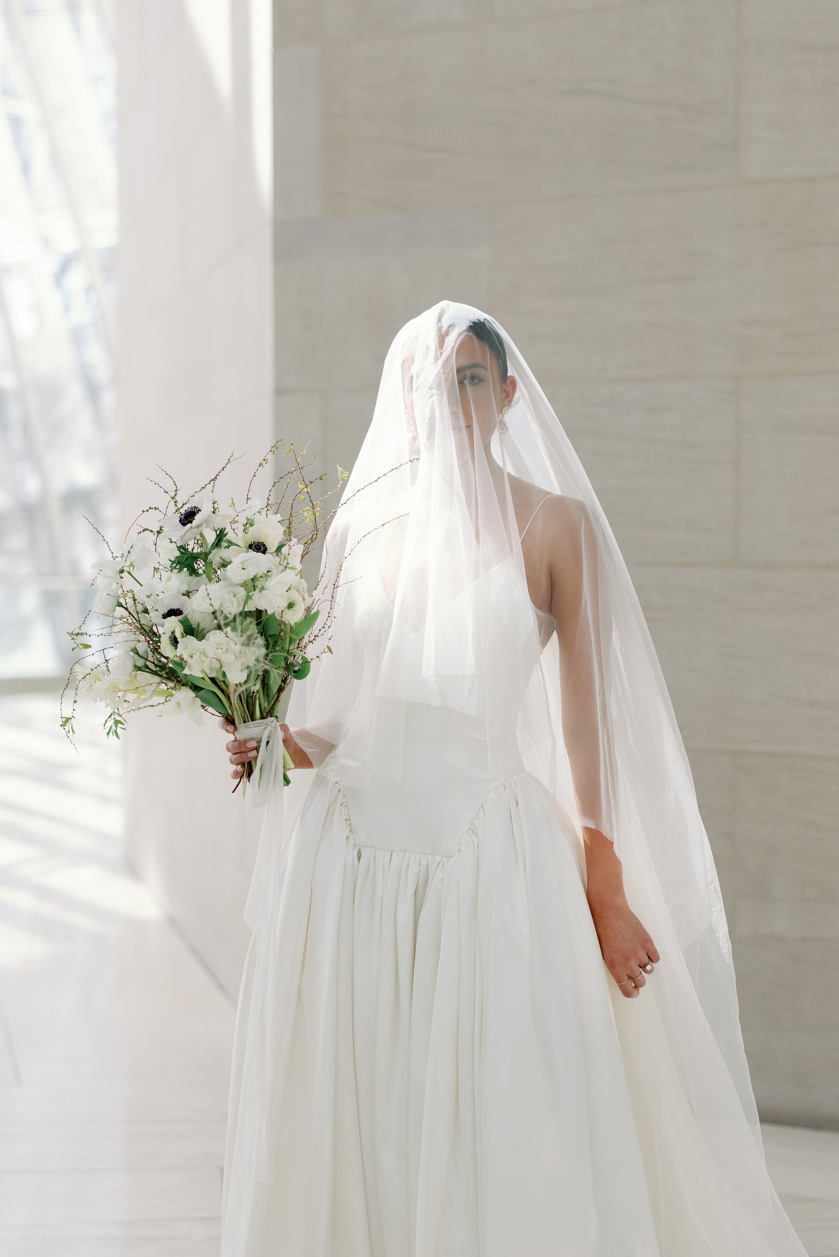 Bride in a white wedding dress with a long veil holding a white neutral tone bouquet in museum.