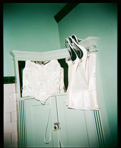 White lace and satin couture bridal corset bustier hanging on a rack against a teal wall.