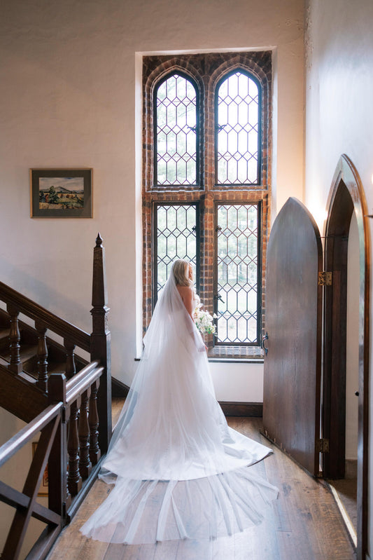 Bride in a white strapless wedding dress standing in a hallway with large stained glass windows and wearing extra long English net royal veil from one blushing bride