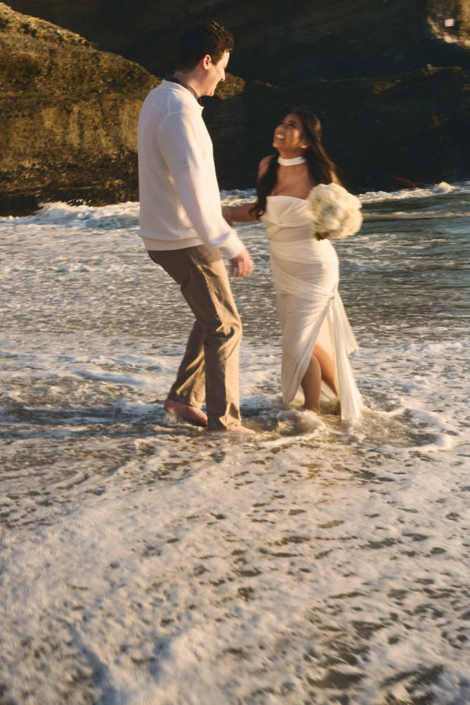 beautiful Mexican bride wearing slit flowy dress in ocean waves and neck scarf