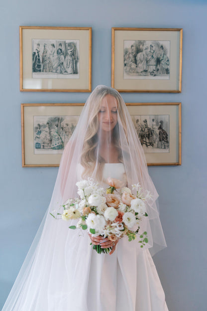 Bride wearing a drop blusher wedding veil holding a bouquet in front of a wall with framed artwork