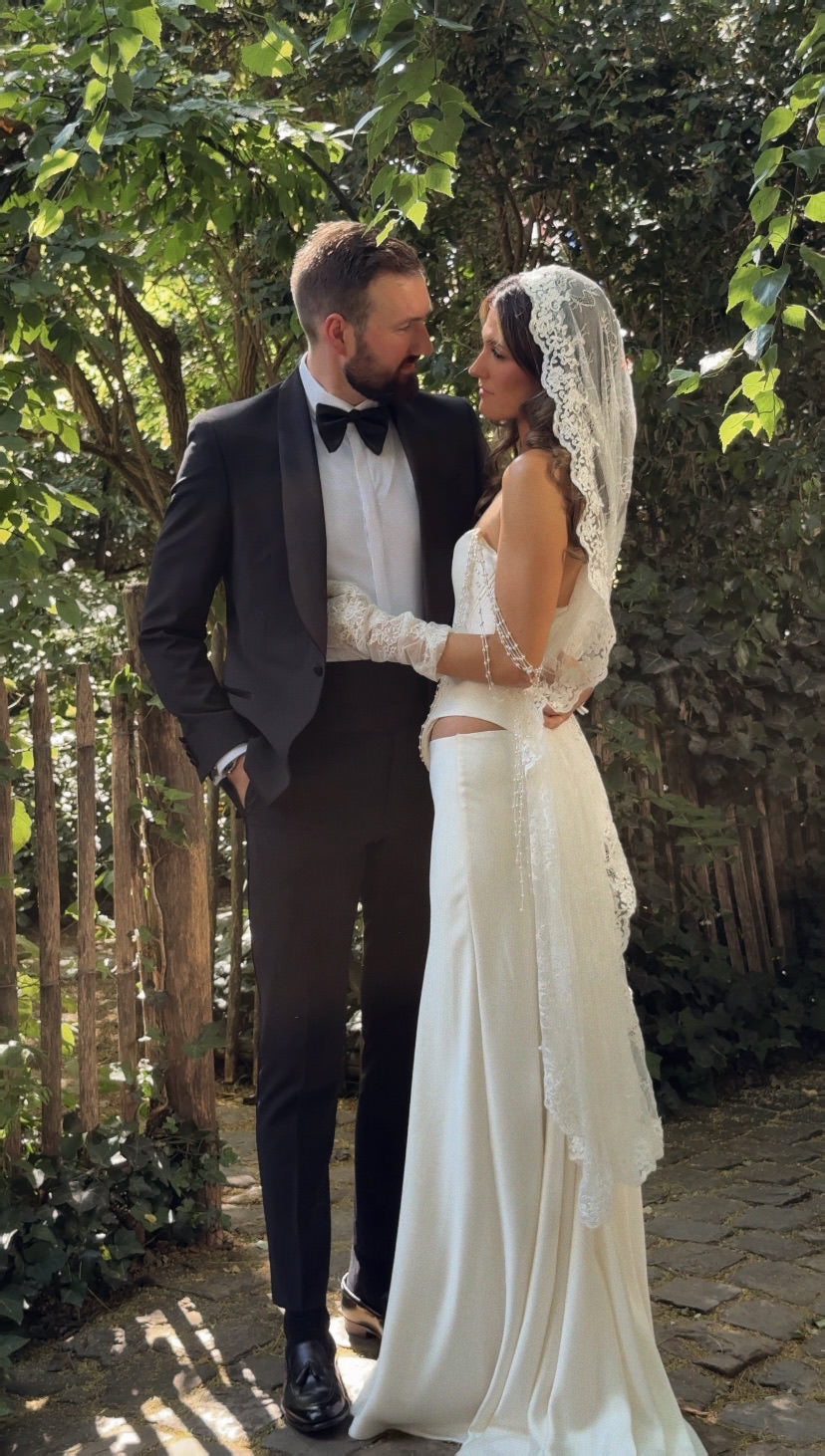 Bride  in vintage inspired  wedding attire and knee length mantilla veil standing outdoors with greenery in the background