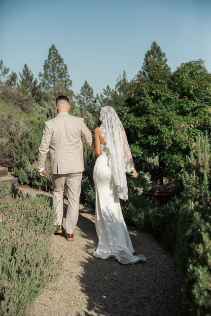 outside garden path with bride walking in knee length lace bridal veil in mantilla style over her head