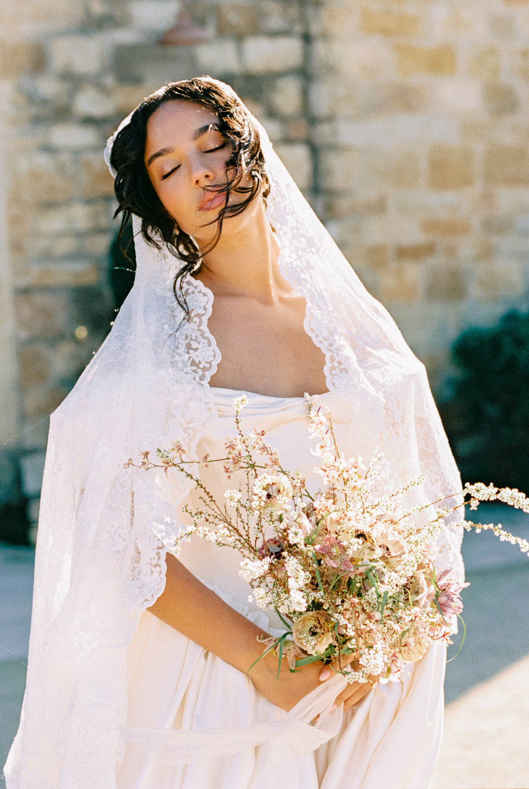 romantic Bride in lace veil holding a bouquet of flowers with a stone wall