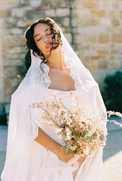 romantic Bride in lace veil holding a bouquet of flowers with a stone wall